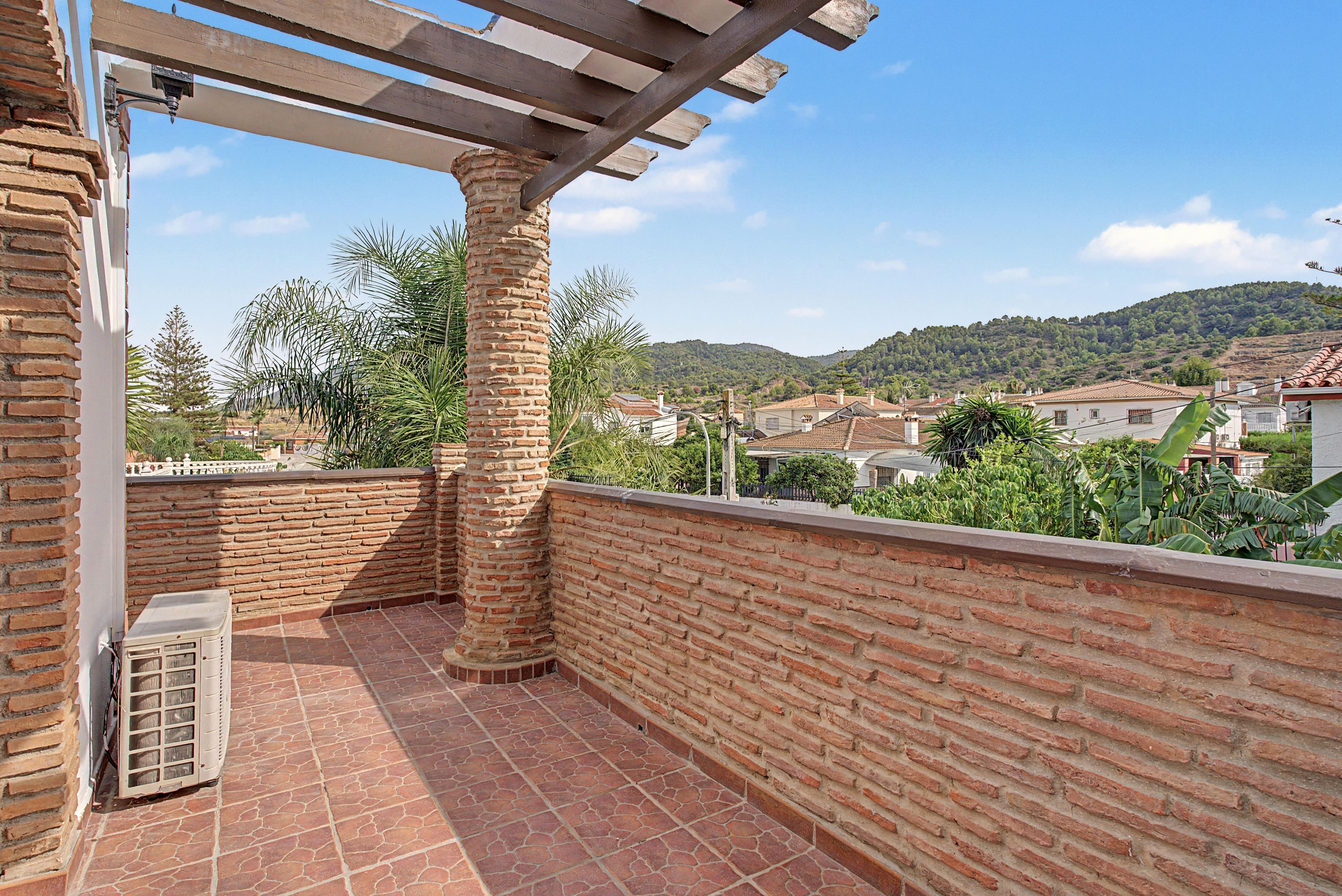 Un balcón con un muro de ladrillo, una pérgola y vistas a una zona residencial con casas y colinas.
