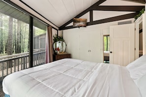 Ein Schlafzimmer mit einem großen Bett, einem Fenster mit Blick auf Bäume, einem Spiegel und einer Tür zu einem anderen Raum.