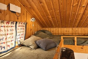 A small, cozy room with wooden walls and a sloped ceiling. There is a bed with a green mattress and two pillows, a small table, and a wall-mounted lamp.