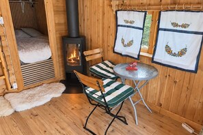 A cozy room with a wood-burning stove, a bed with a white blanket, two striped chairs, a small round table, and a window with curtains.