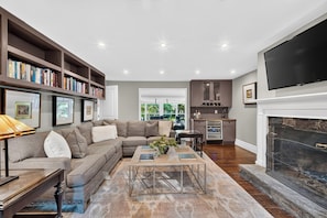 A living room with a sectional sofa, a coffee table, a bookshelf, a fireplace, and a kitchen area. - Private Pool + Swim + Fish the Lake + Kayak | Waccabuc Lake House (Waccabuc)