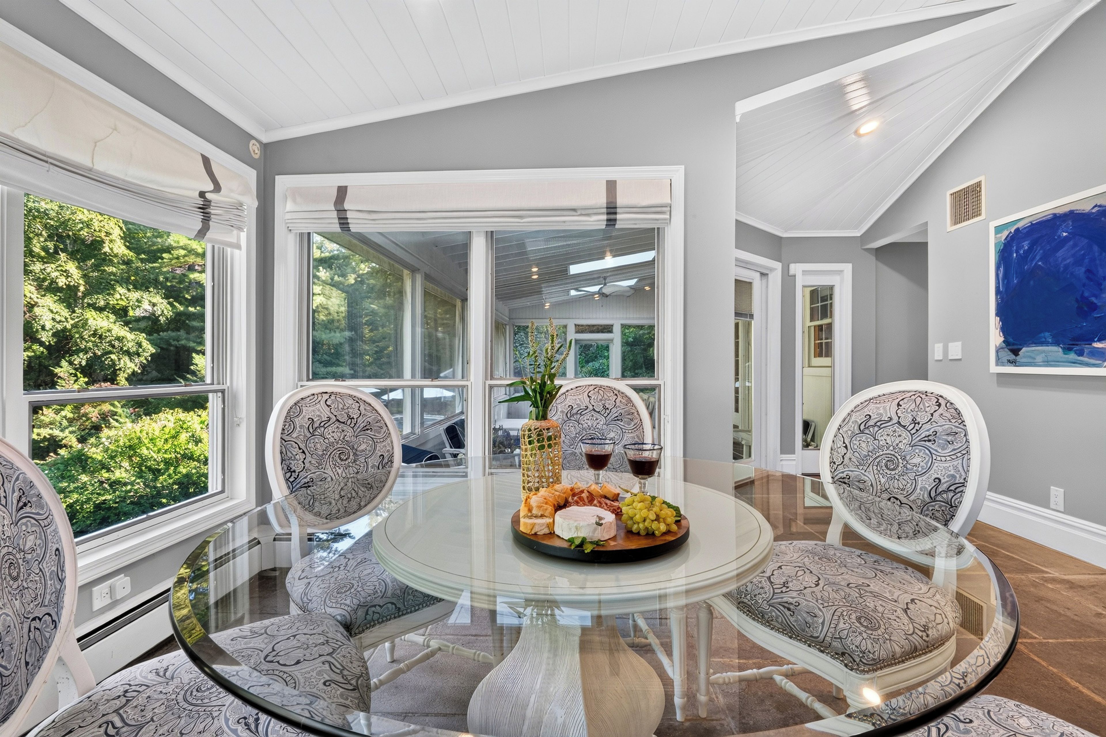 A dining area with a glass table surrounded by patterned chairs, a centerpiece with cheese and grapes, and a view of greenery through large windows.