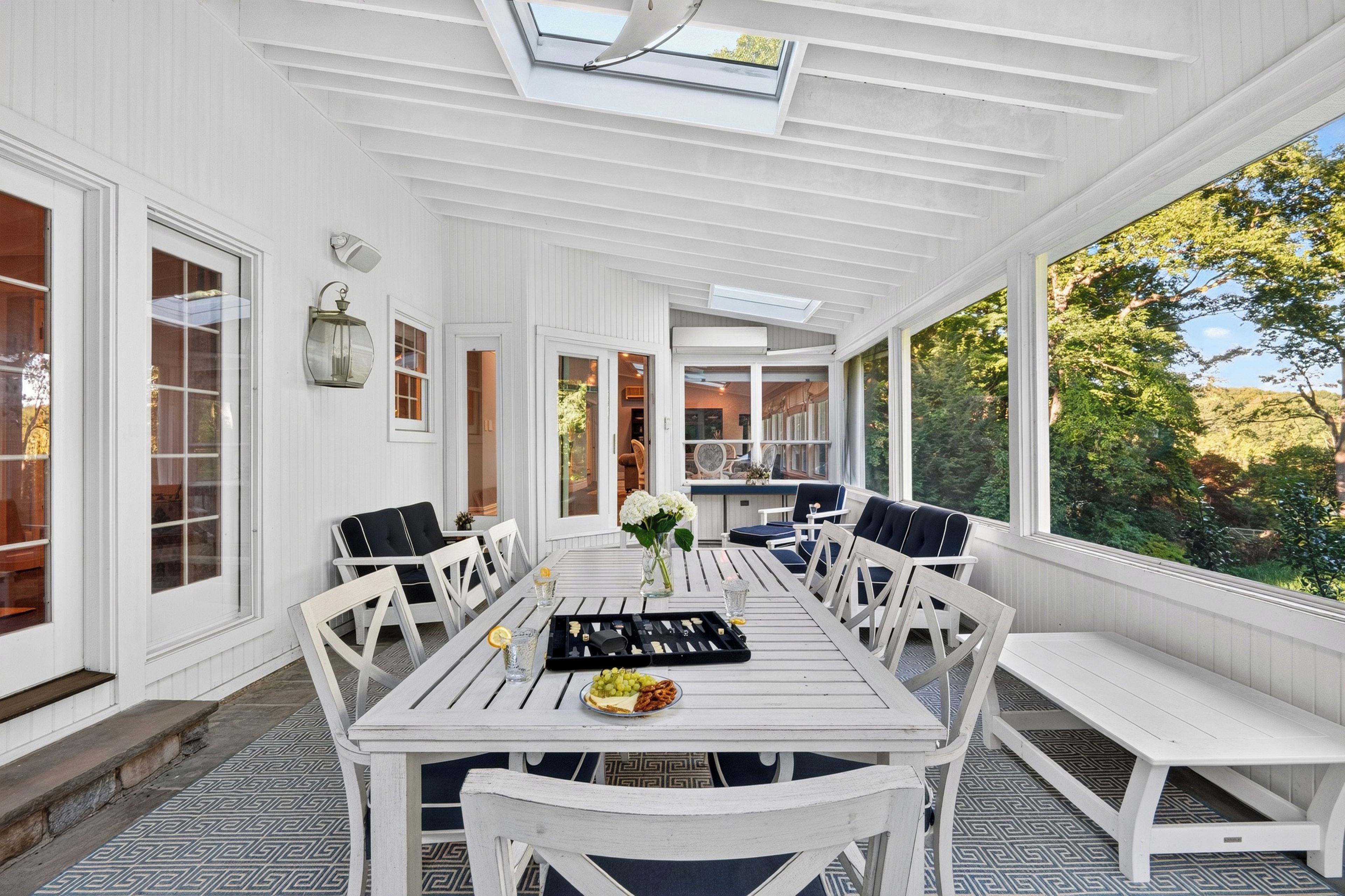 A covered patio with a white table set for dining, surrounded by white chairs, and a bench.