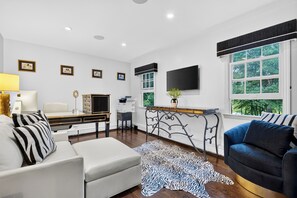 A modern living room with a sofa, a coffee table, a TV mounted on the wall, and a window with a view of trees. - Private Pool + Swim + Fish the Lake + Kayak | Waccabuc Lake House (Waccabuc)