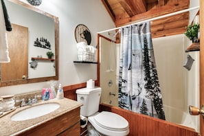 Ein Badezimmer mit einem Holzbadezimmerschrank, einem Waschbecken, einer Toilette, einem Duschvorhang mit Waldmuster und einem Ablagebereich für Pflegeprodukte.