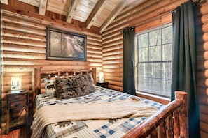 Ein Schlafzimmer in einer Blockhütte mit einem Bett, einem Nachttisch, einem gerahmten Bild an der Wand und einem Fenster mit Vorhängen.