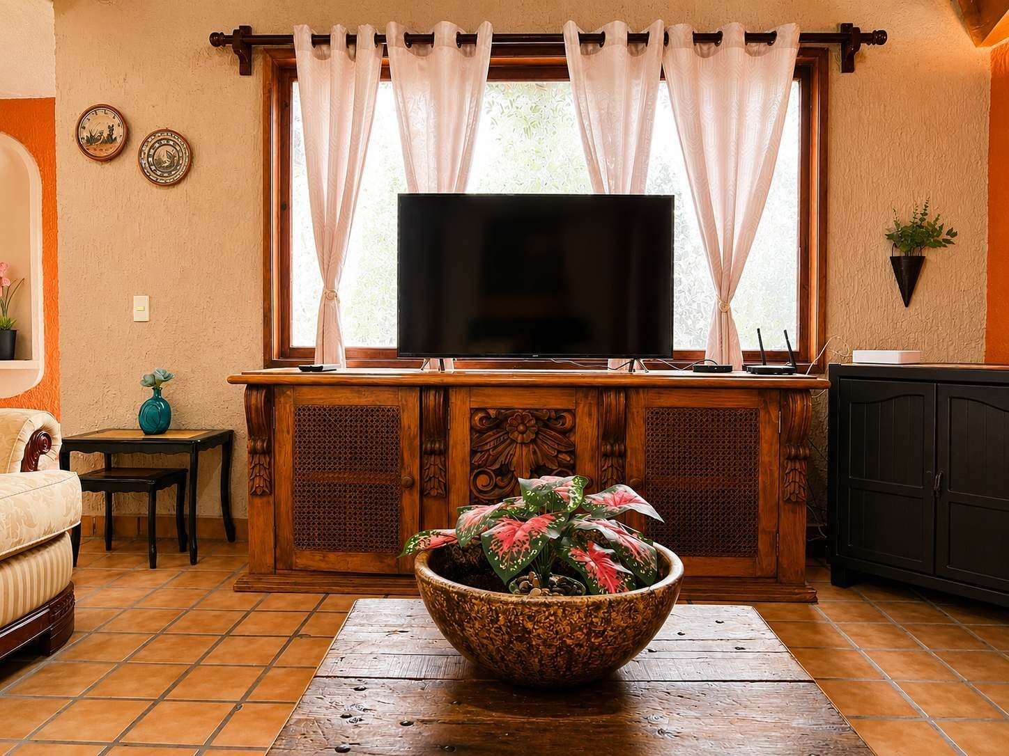 Una sala de estar con televisor, un mueble de madera, un tazón decorativo grande y una ventana con cortinas.
