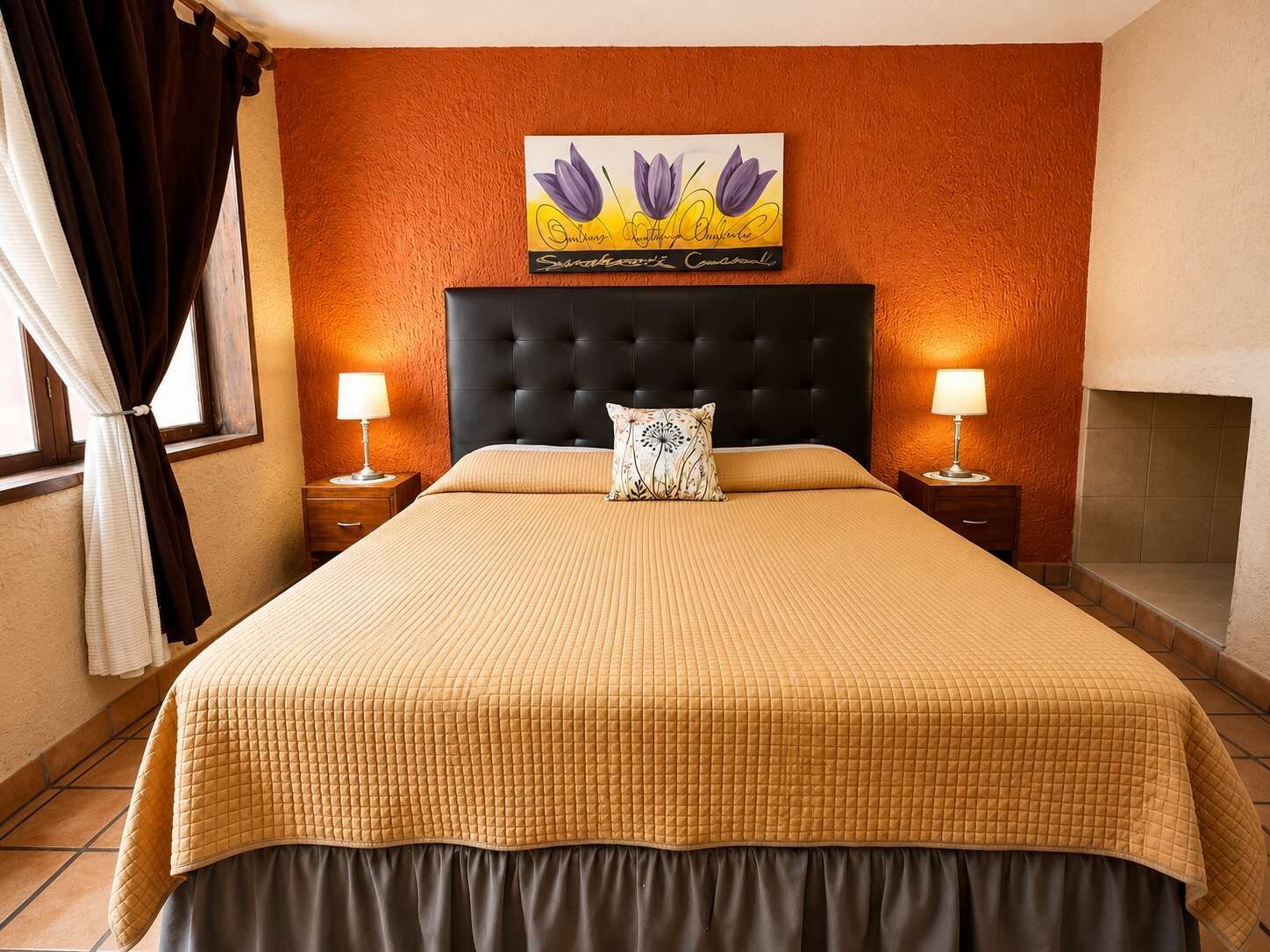 Habitación de hotel con una cama grande, dos mesitas de noche con lámparas, una chimenea y un cuadro floral en la pared.