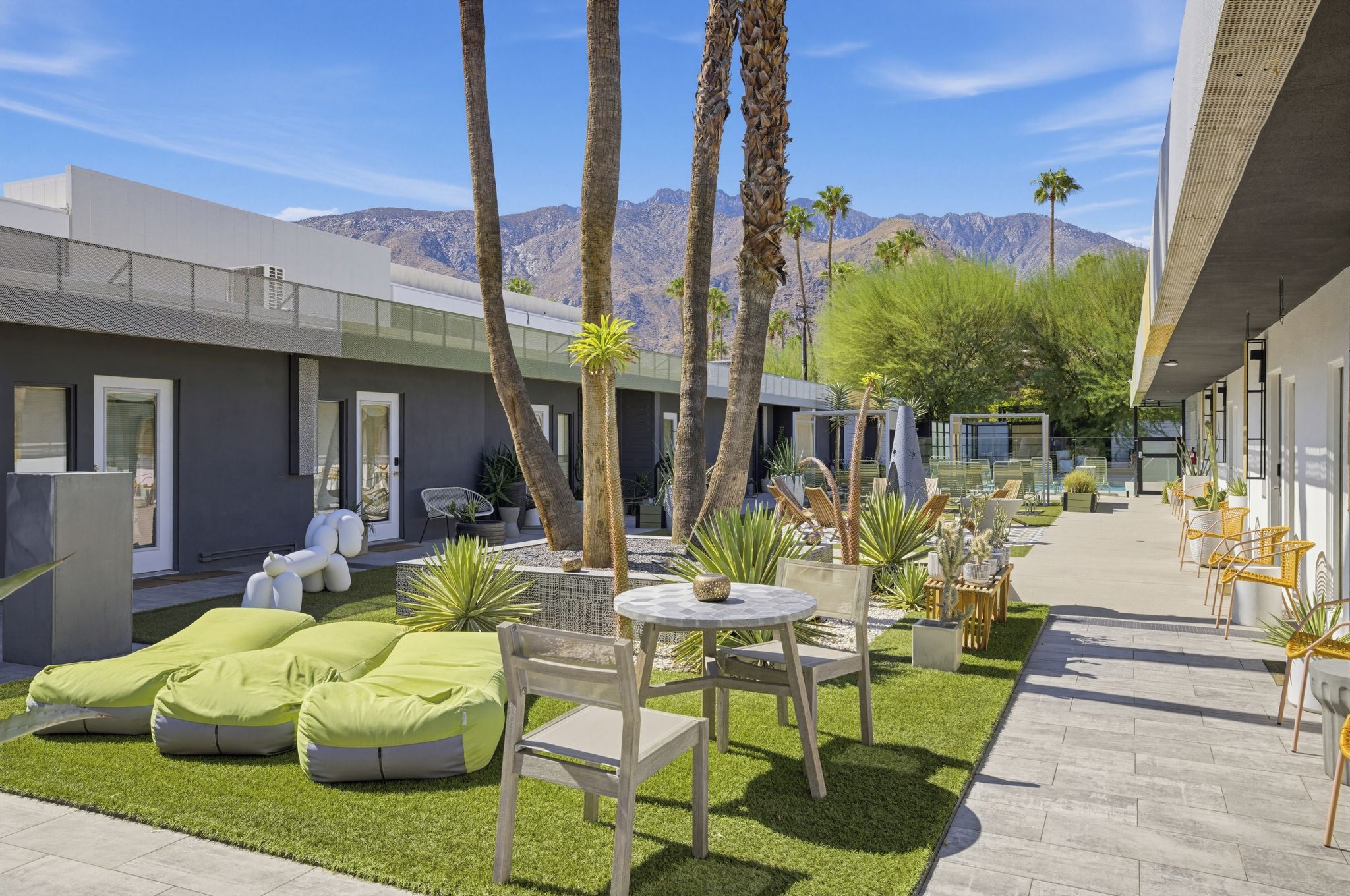 A modern outdoor seating area with green bean bags and tables, surrounded by palm trees and a mountain view.