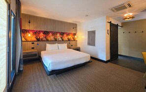 A hotel room with a bed, a headboard with a painting, a wall-mounted air conditioner, and a sliding door. - Semi-Private Patio Facing Pool + Firepit | Mtn Views | Garden King at Mahala (Palm Springs)