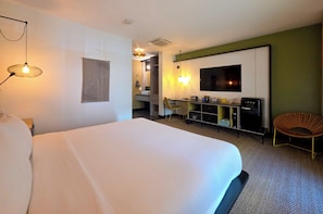 Habitación de hotel con una cama grande, televisor montado en la pared, un escritorio con silla y un pequeño rincón de descanso con una butaca.