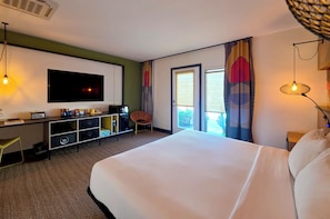 Habitación de hotel con una cama grande, un escritorio con televisor de pantalla plana, una silla, un mueble pequeño y una ventana con cortinas.