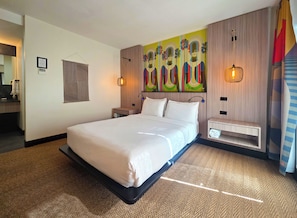 Quarto de hotel moderno com uma cama grande, cabeceira com um mural colorido, mesa de cabeceira com abajur e uma pequena estante.