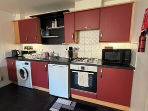 A compact kitchen with red cabinets, a washing machine, a dishwasher, a microwave, and a gas stove. - A&R Guest House - 2-Bed Coach House, Bristol (Bradley Stoke)