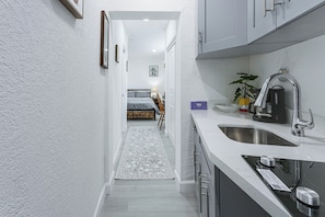 A narrow hallway leading to a bedroom with a bed, a bedside table, and a lamp. - Chic North Miami Studio - 6 Mi to Beach! (North Miami)