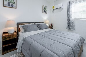 A neatly made bed with a grey quilt, a bedside lamp, and a nightstand with a book and a small lamp.