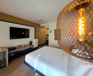 A hotel room with a bed, a TV mounted on the wall, a small refrigerator, and a decorative light fixture. - Pool + Spa + Firepit + Courtyard | Mtn Views | Sun Queen at Mahala (Palm Springs)