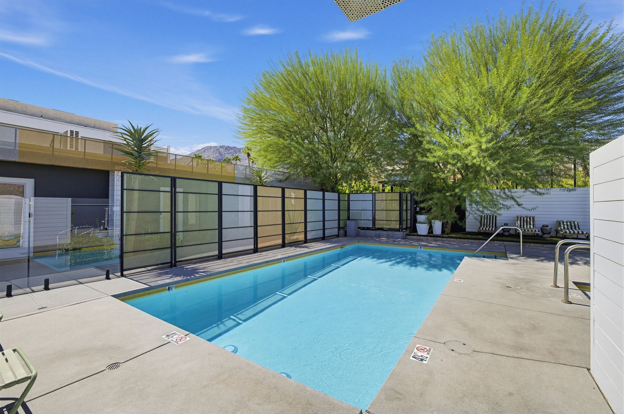 Una piscina rectangular con superficie de agua azul cristalina, rodeada por un borde de hormigón y una valla de metal.