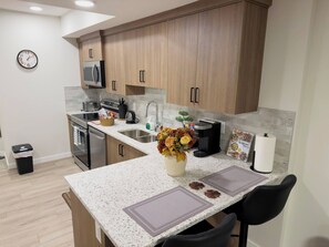 A modern kitchen with wooden cabinets, stainless steel appliances, a granite countertop, and a small dining area with a chair. - Private Suite Near Calgary Airport (Calgary)