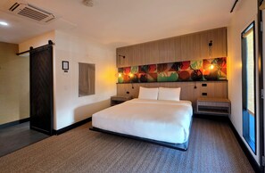 A hotel room with a large bed, a wooden headboard, a colorful artwork above the bed, a small bedside table, and a large window. - Pool + Spa + Firepit + Courtyard | Mtn Views | Desert King at Mahala (Palm Springs)