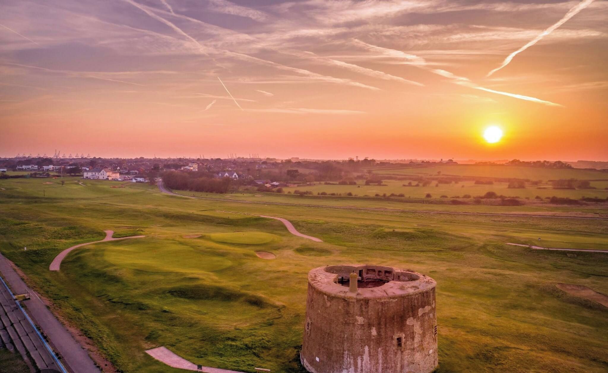 A grassy field with a circular structure in the foreground and a sunset in the background.