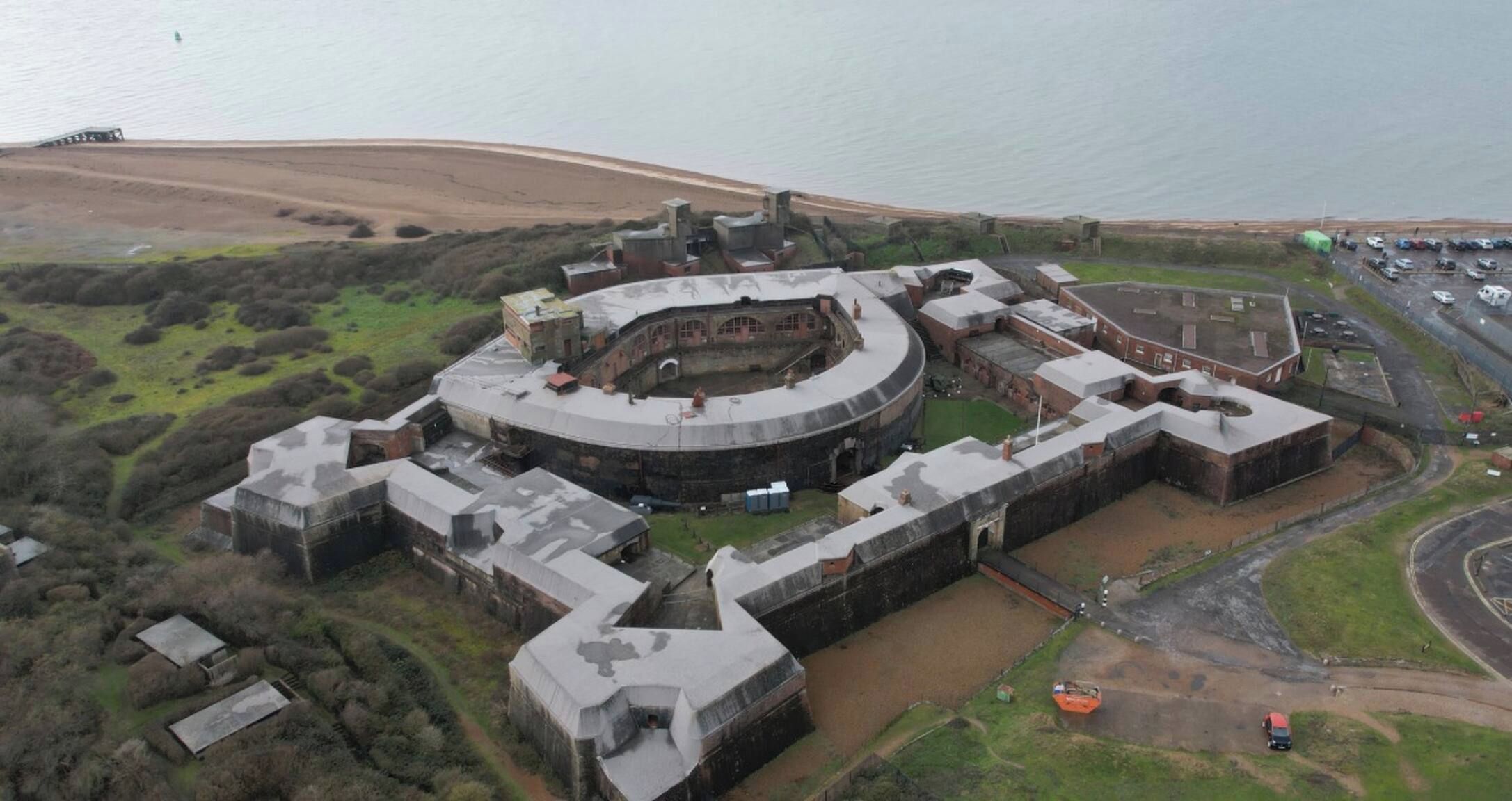 Un grand bâtiment circulaire, doté de plusieurs étages et d’une cour intérieure, entouré de vastes champs et d’une étendue d’eau.