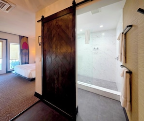 Habitación de hotel con cama, puerta corredera, zona de ducha con mampara de vidrio y toallero.