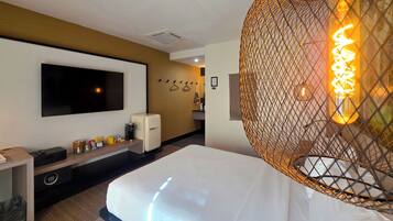 A hotel room with a bed, a TV mounted on the wall, a small refrigerator, and a decorative light fixture.