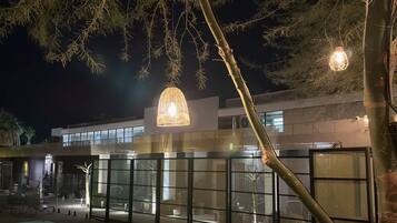 A swimming pool area at night with a modern building in the background, surrounded by a fence and illuminated by hanging lights.