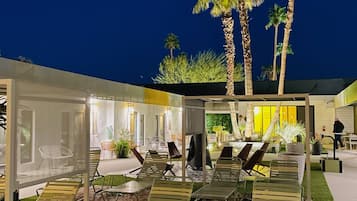 A well-lit outdoor lounge area with sun loungers and chairs, surrounded by palm trees and a covered seating area.