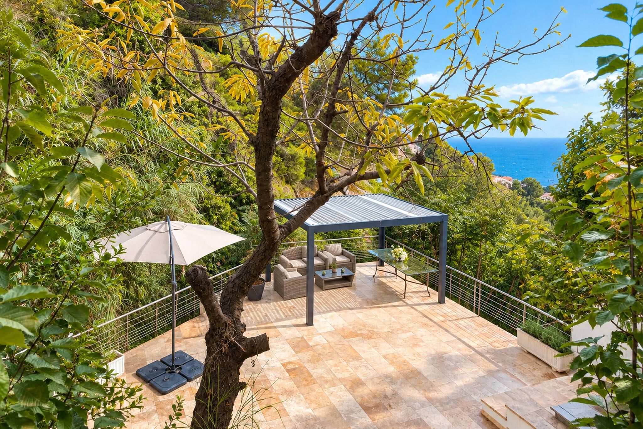 Un espace extérieur aménagé, abrité et doté de tables et de chaises, entouré d’arbres et offrant une vue sur une étendue d’eau.