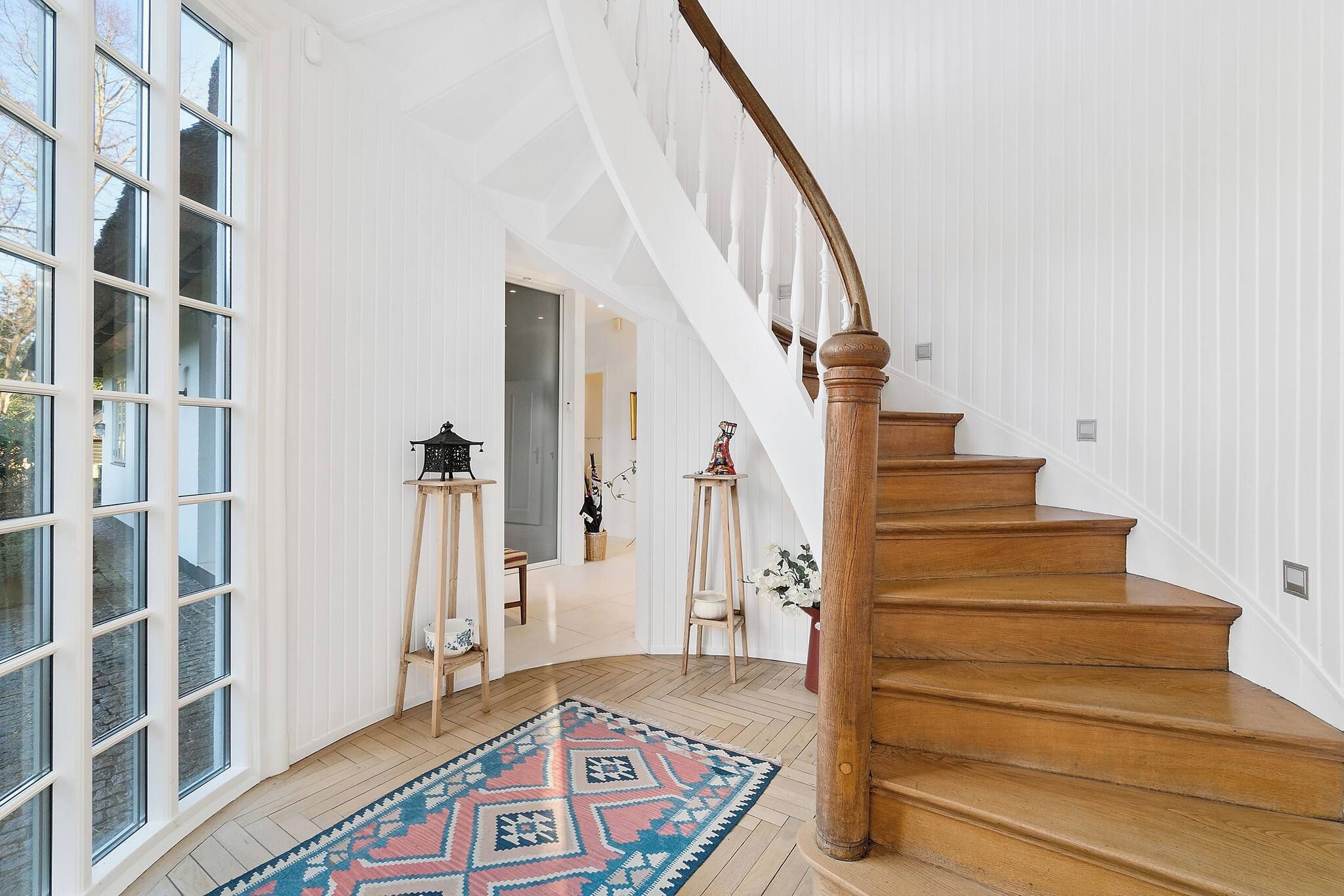 A spacious interior with a wooden staircase, large windows, and a patterned rug.