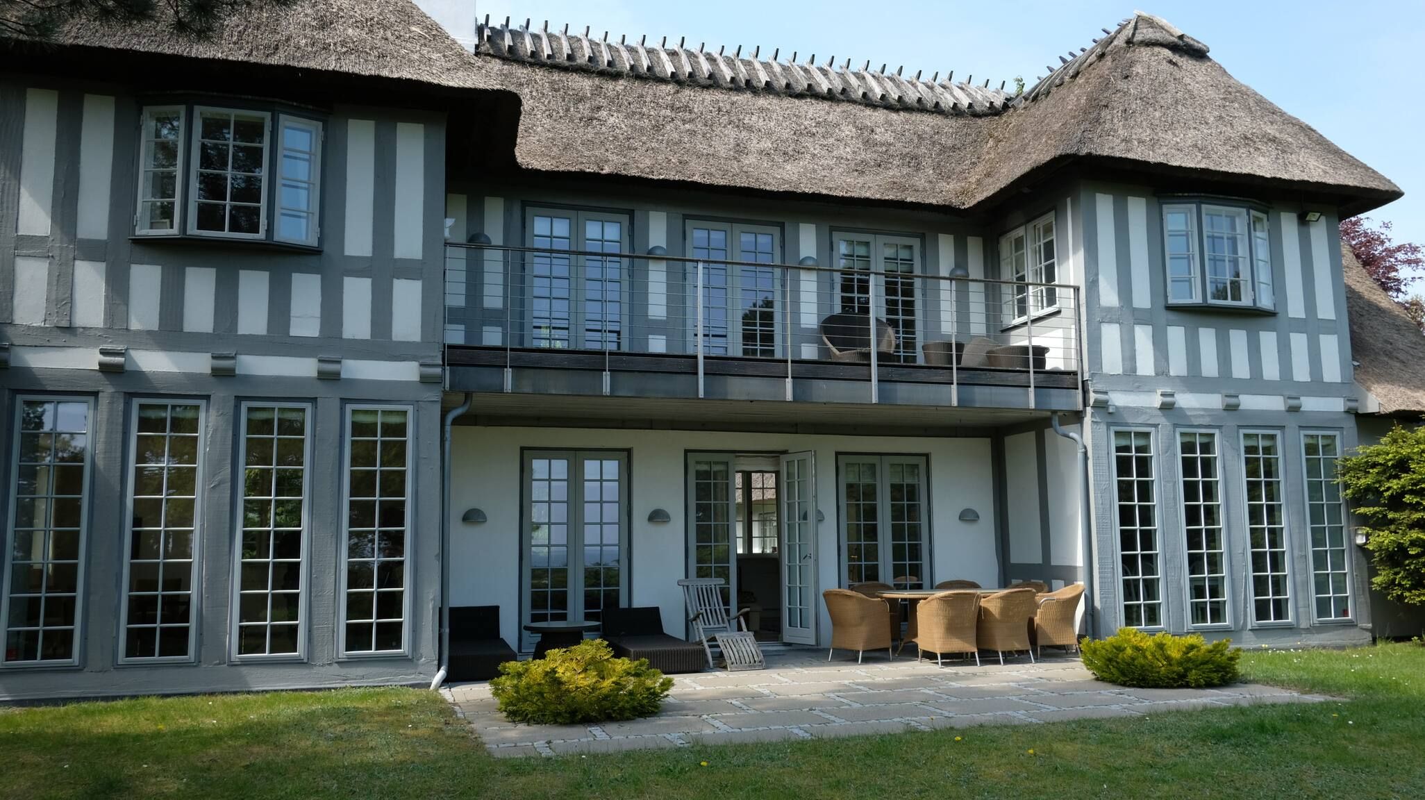 Una casa de dos pisos con techo de paja, ventanas grandes y un balcón con mobiliario exterior.