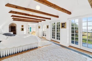 A spacious bedroom with a large bed, wooden beams, white walls, and a view of the outdoors.