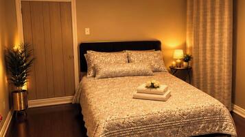 Una cama bien hecha con una colcha y almohadas estampadas, una lámpara de noche, una mesita con una planta y una puerta con panel de madera.