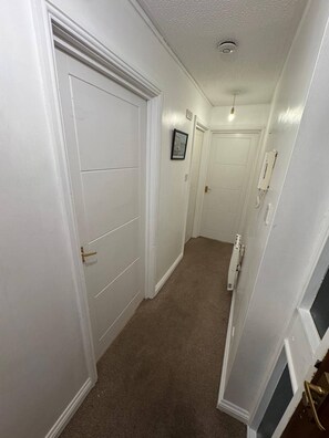 A narrow hallway with white walls and carpeted flooring, leading to a door with a brass handle. - Charming 1-Bedroom Apartment in West Northamptonshire (West Northamptonshire)