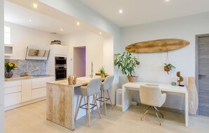 Una cocina moderna con armarios blancos, una isla de madera y un comedor con mesa y sillas blancas.