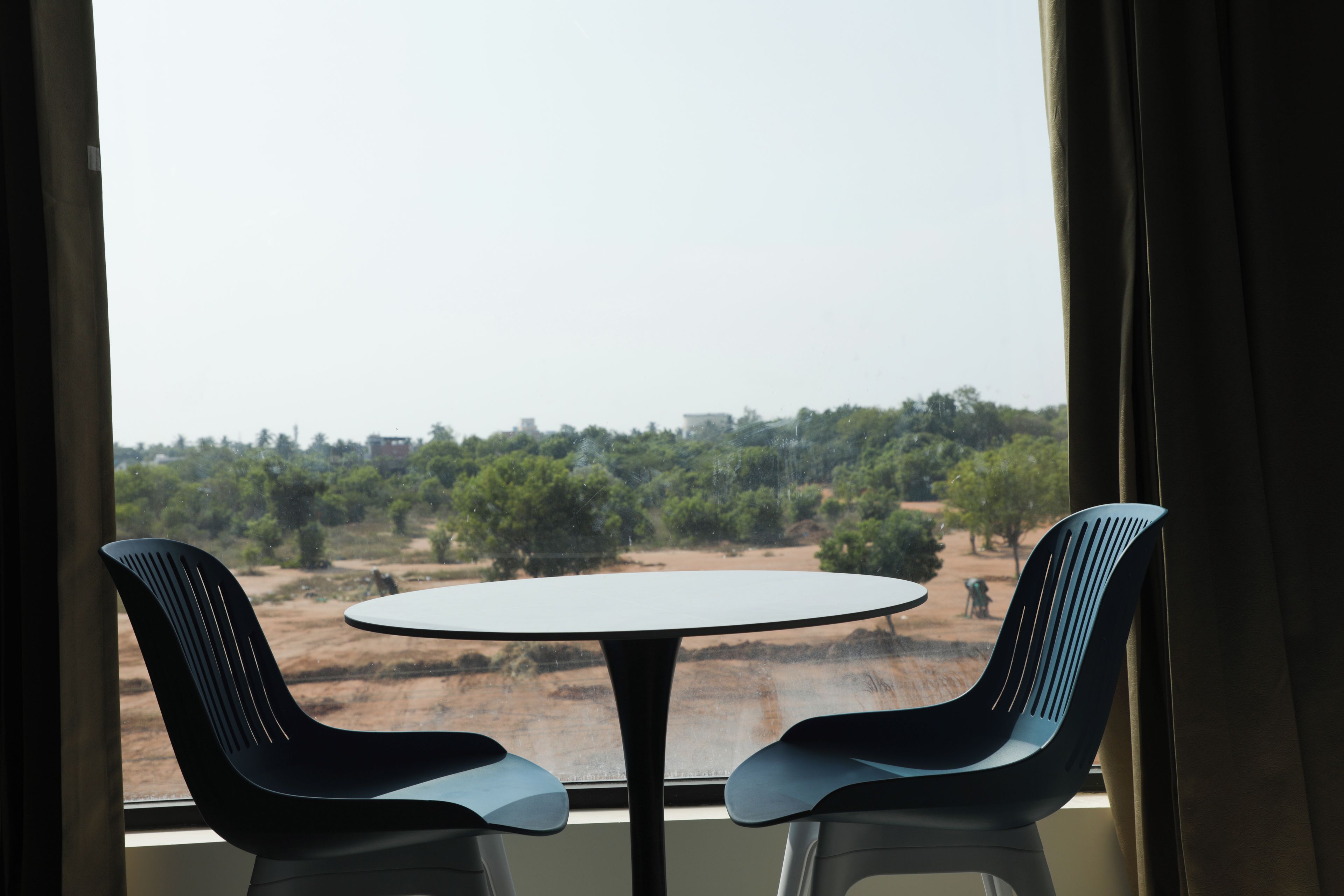 Quarto com mesa redonda e duas cadeiras, janela com cortinas e vista para árvores e edifícios.