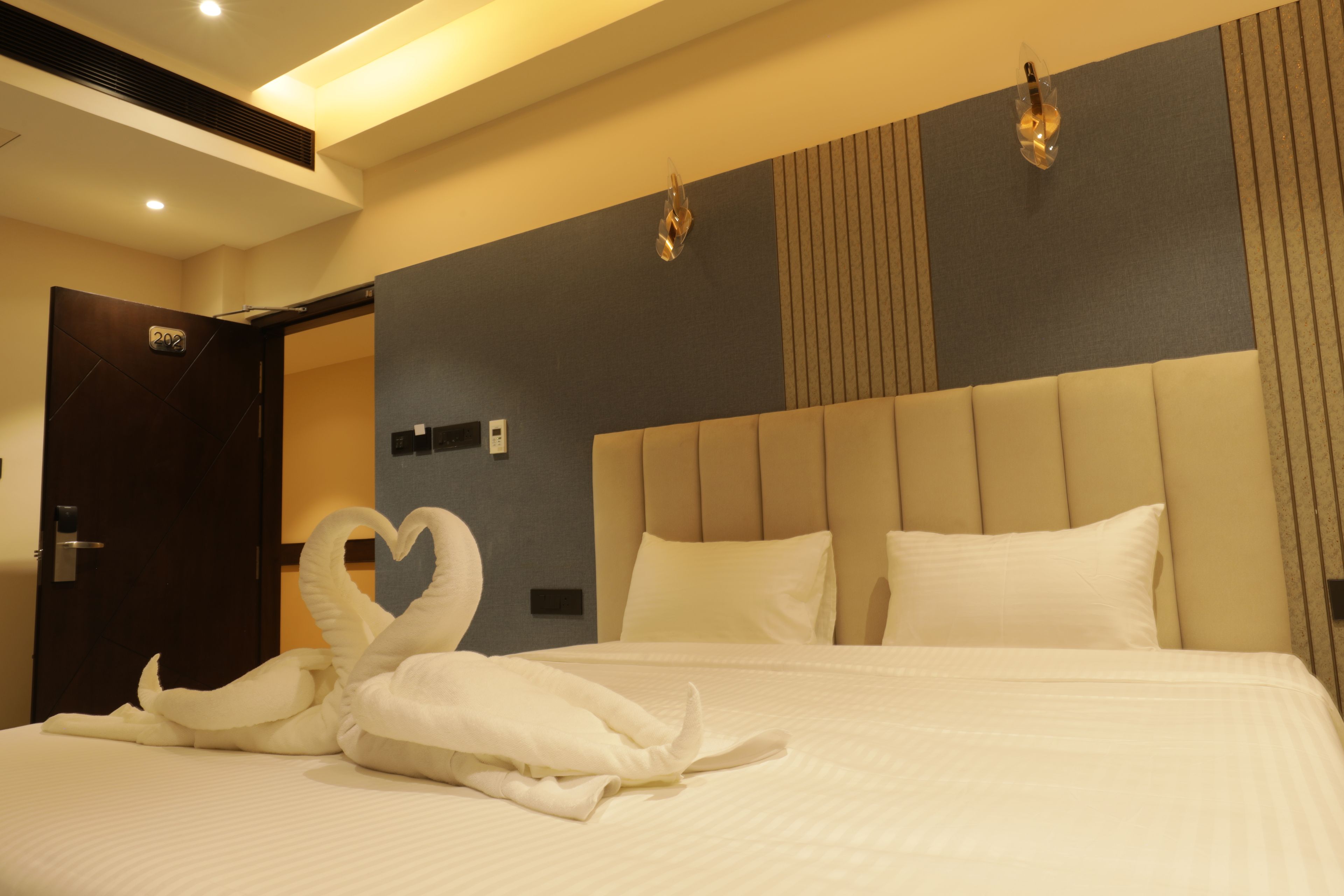 A hotel room with a bed, a heart-shaped towel arrangement, a door, and a wall with a light fixture.