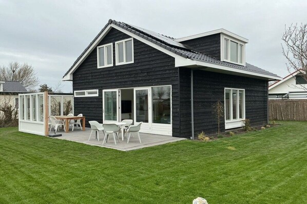 Una casa moderna de dos pisos con fachada oscura, ventanas amplias y un patio con mobiliario exterior.