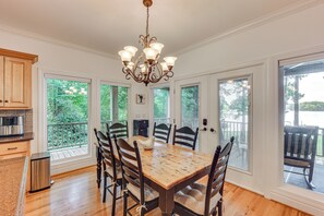 A dining area with a wooden table and chairs, a chandelier, and large windows with a view of greenery. - Dock, Hot Tub, Game Room! Lake Wateree Retreat (Camden)