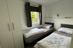 A room with two single beds, a wardrobe, a window with curtains, and a small shelf with items.
