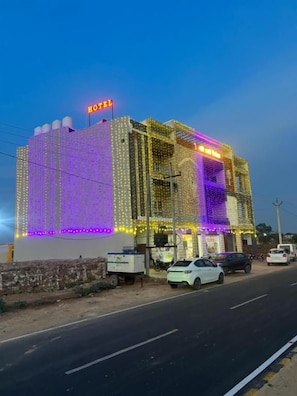 A building with a 'HOTEL' sign, multiple floors, and a parking area with cars. - SHREE RADHE PALACE (Degana)