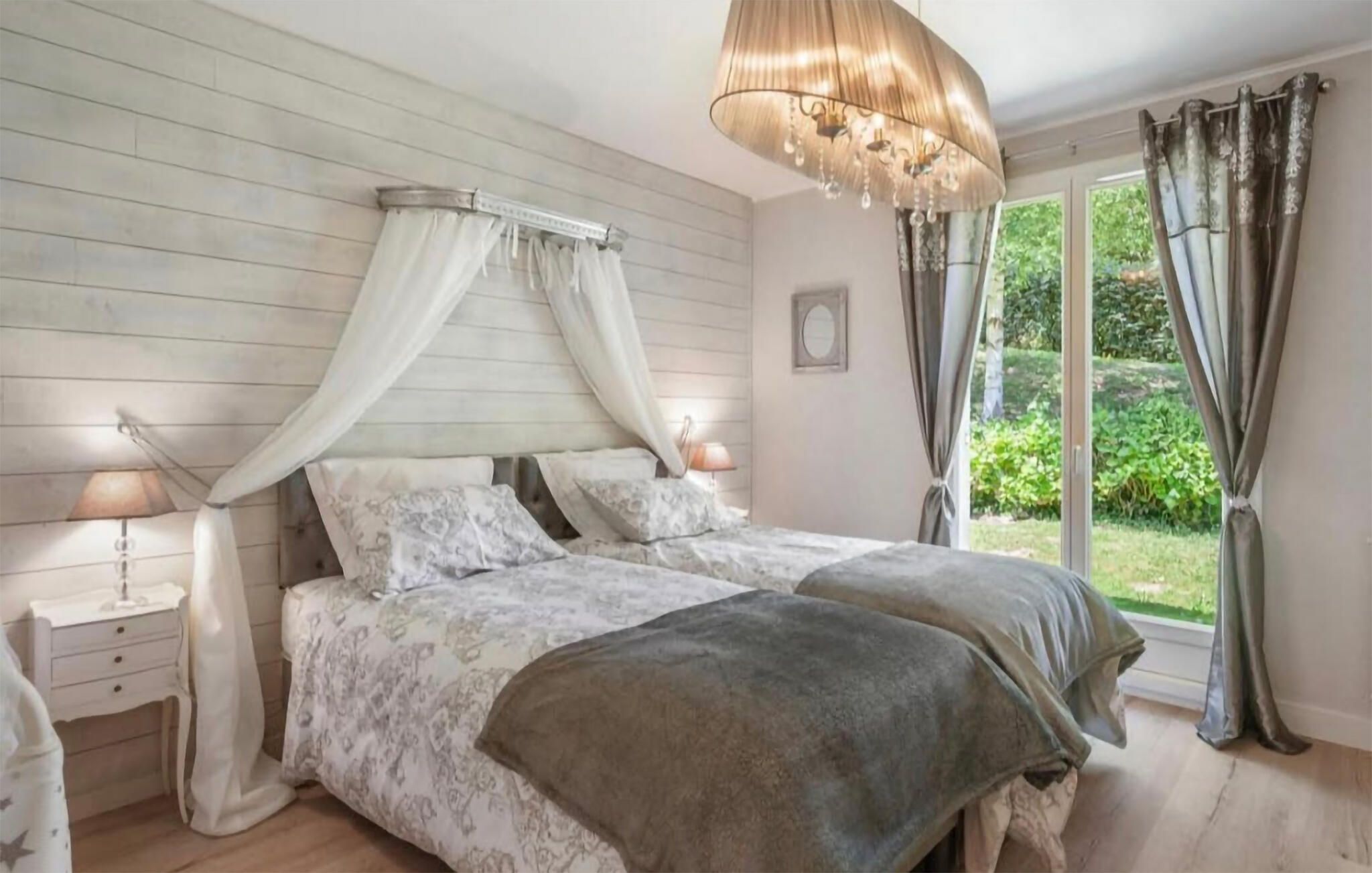 A bedroom with a canopy bed, bedside lamps, a large window with curtains, and a chandelier.
