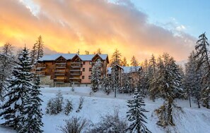 Ein mehrstöckiges Blockhaus mit Balkonen, umgeben von schneebedeckten Bäumen, vor einem Sonnenuntergangshimmel.