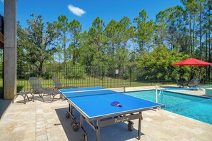 An outdoor table tennis table with a net, chairs, and a pool in the background. - Play & Unwind! Pass Christian Home < 1 Mi to Gulf (Pass Christian)