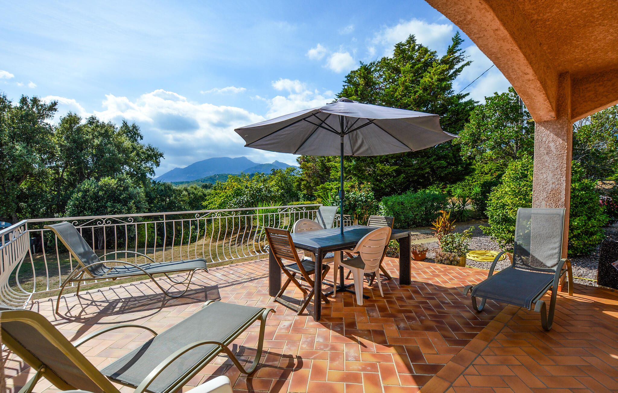 An outdoor patio with a table and chairs, a large umbrella, and a view of a mountainous landscape.