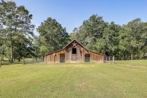 A rustic wooden barn with a star on the roof, surrounded by a fenced pasture and tall trees.