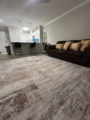 A living room with a brown couch, a patterned rug, and a kitchen in the background.