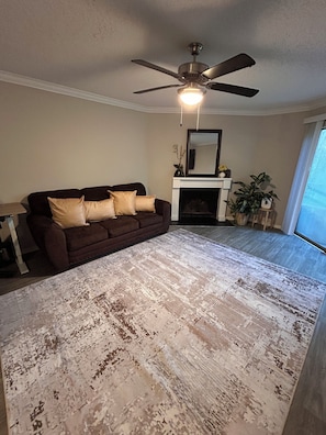 A living room with a brown sofa, a fireplace, a ceiling fan, and a large window.